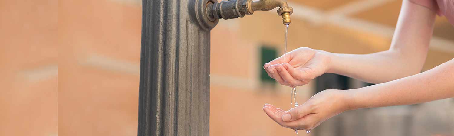 Serving Wastewater Issues Tirelessly 1 Water slowly drips from an old outdoor faucet into cupped hands, symbolizing water scarcity and wastewater issues.