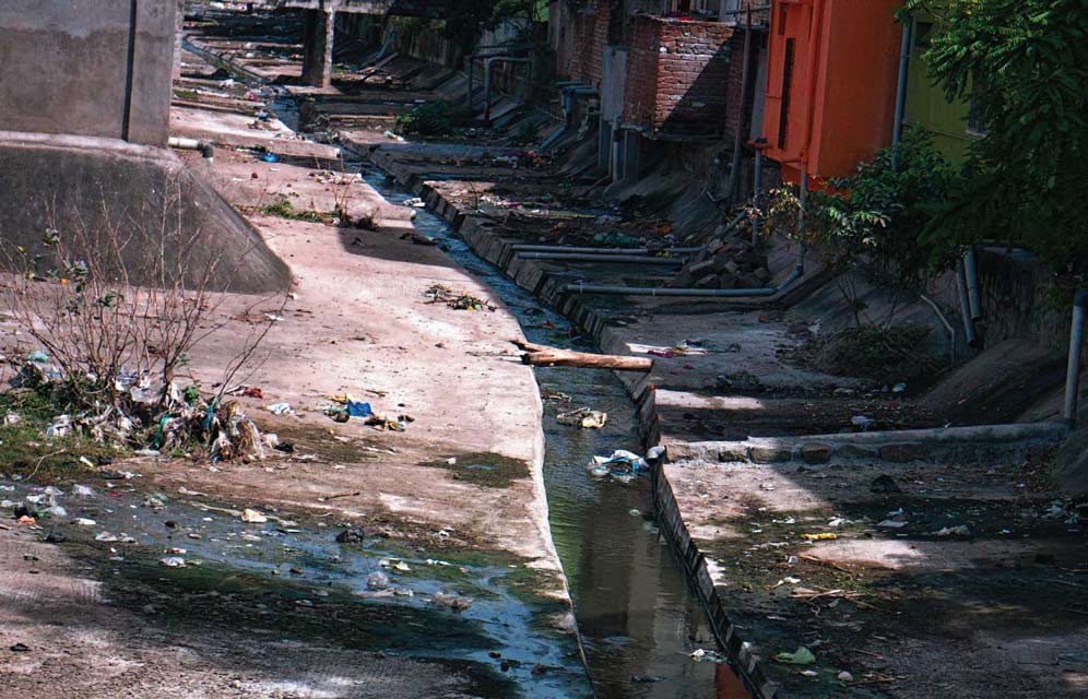 Monsoons & Mumbai: How Diseases Spread Due To Ineffective Sanitation 1 A narrow, open drainage channel filled with stagnant, polluted water and garbage next to buildings, typical of Mumbai's sanitation issues during monsoons.