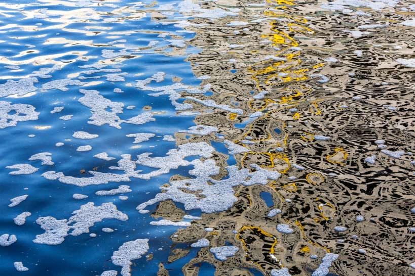 Our Oceans – Every Drop Counts 1 A close-up of ocean water with white foam on one side and reflections of yellow and dark elements on the other, highlighting pollution and the importance of every drop.
