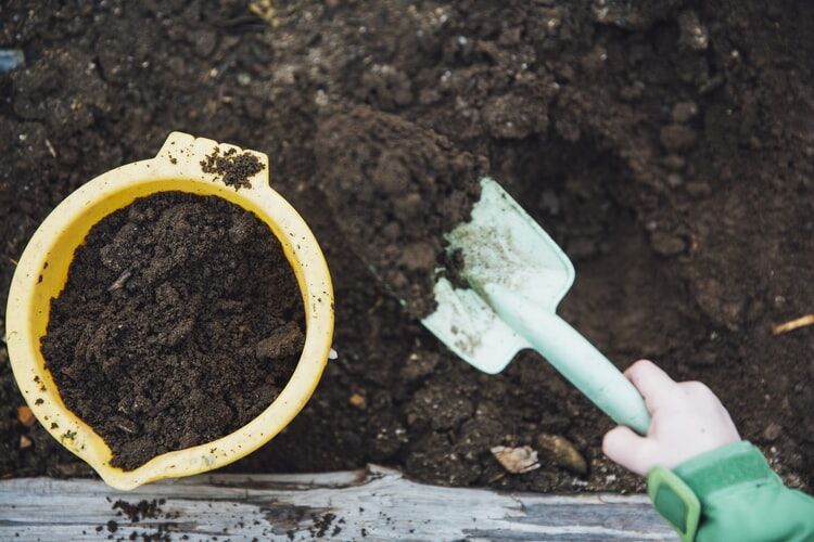 Your Complete Guide To Home Composting 1 A child's hand with a small shovel next to a yellow bucket full of rich, dark compost, representing home composting.