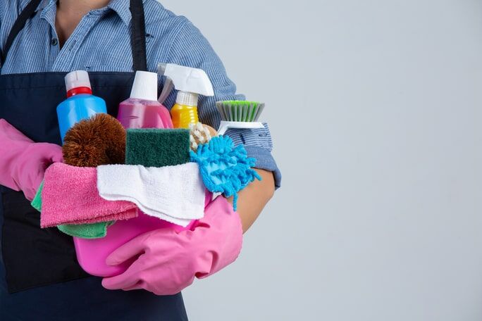 4 Types of Chemical Cleaning Agents and Why Not to Use Them 1 A person wearing pink gloves and an apron holds a caddy filled with various colorful chemical cleaning agents.