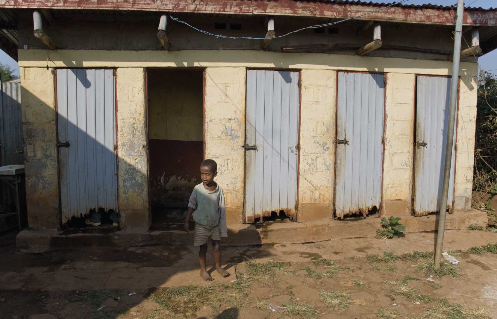 The Pressing Issue Of Sanitation In Nigeria 1 A young child stands barefoot in front of a row of basic, weathered public latrines, depicting sanitation conditions in rural Nigeria.