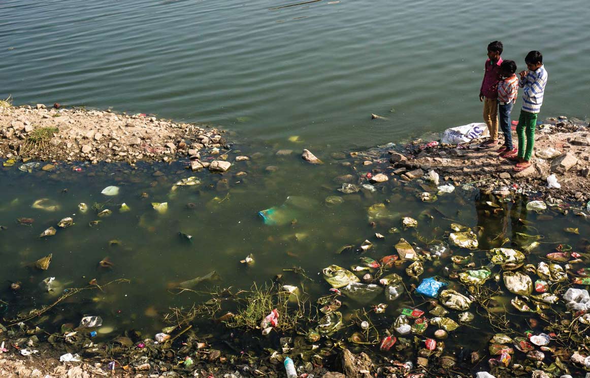 Sanitation And The Unlikely Link To Good Health 1 Three children stand by a highly polluted body of water filled with trash, highlighting poor sanitation and health hazards.