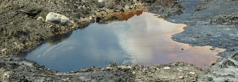 How To Remediate Oil Contaminated Soil 1 Oil-contaminated soil with a puddle reflecting the sky, surrounded by dark, polluted ground and rocks.