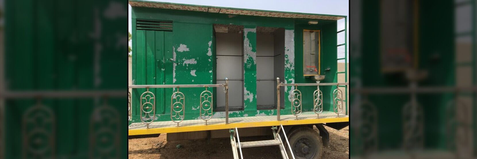 A green, weathered municipal mobile toilet unit with multiple stalls and a railing, suitable for public sanitation needs.